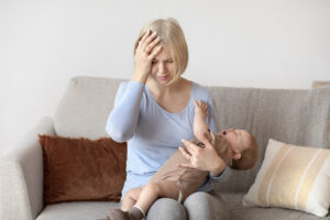 Exhausted blonde woman young mother having migraine, holding crying little baby, home interior, copy space. Tired caucasian mom having headache, touching her forehead. Stressful motherhood concept